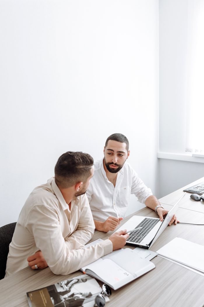 gallery-05 Two businessmen in white shirts engaged in a discussion over a laptop in a bright office setting.